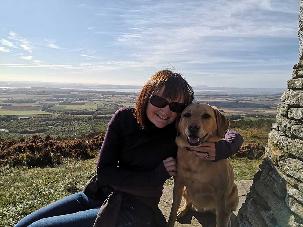Jenny and her dog photographed at the top of a hill next to a cairn on a summers day. 